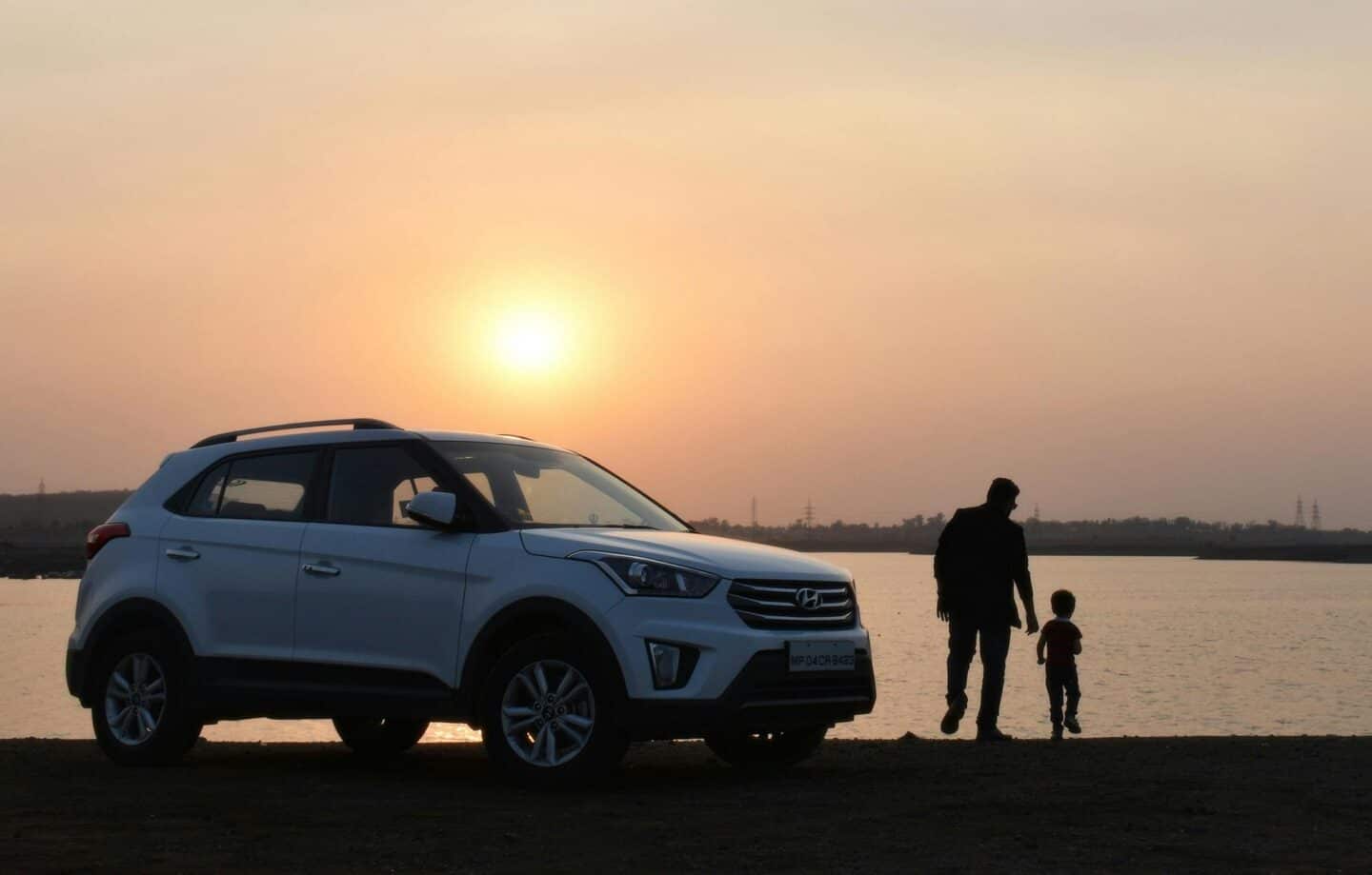 Man and child stood next to a white car by a lake at sunset